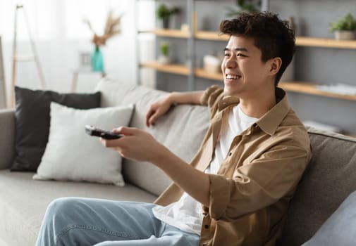 Leisure And Entertainment. Smiling asian guy watching TV show or film, holding remote control. Young man enjoying free time sitting on the comfortable couch at home in living room, free space