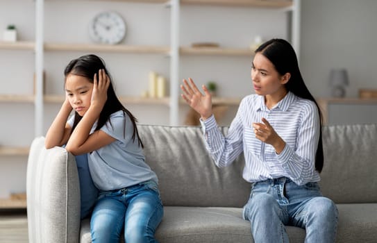 Mother daughter conflict. Young korean mother trying to speak to her upset daugter, sitting on sofa at home. Offended asian girl covering her ears, not wishing to listen to parent