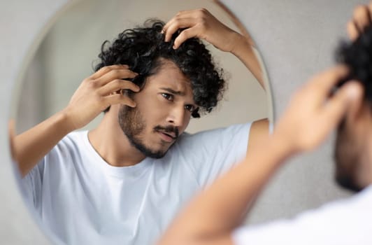 Men's beauty and haircare treatment. Worried indian guy looking at flaky scalp in mirror, examining gray hair and hairloss issue, standing in bathroom at home.