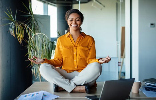 Office zen. Calm black businesswoman meditating with closed eyes on office desk, sitting at workplace in lotus position. Positive woman feeling peaceful and balanced
