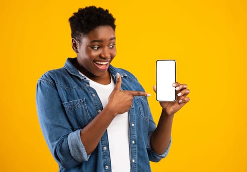 Cheerful Black Lady Showing Smartphone Blank Screen Recommending Application Posing Standing Over Yellow Studio Background. Mobile Application Ad. Check This App Concept. Mockup