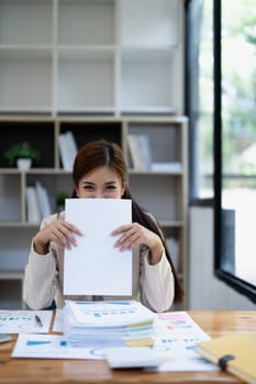 Young woman in beige long-sleeved shirt playfully hiding her face with a blank sheet at a desk cluttered with heaps of paperwork and documents..