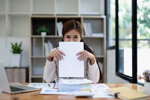 Young smiling woman playfully hiding behind large blank paper sheet at modern office desk with documents stacked in front of her for creative concept.