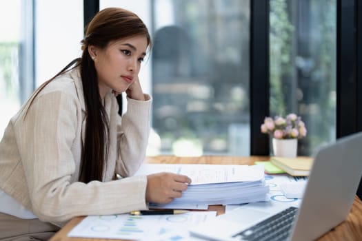 Stressed young businesswoman overwhelmed by paperwork with finance, market charts and documents in office.