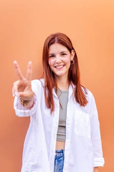 young woman standing in front of orange wall holding up two fingers in victory sign and smiling looking at camera, concept of peace, love and positivity, copyspace for text