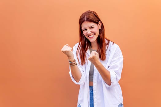young woman standing in front of an orange wall celebrating with clenched fists and smiling very happy, concept of concept of success and happiness