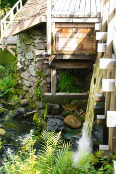 Old watermill surrounded by green nature. A small stream flows over moss-covered stones, surrounded by trees. Idyllic landscape with rustic wooden architecture.