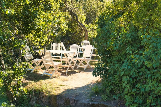 A white garden furniture set stands idyllically in the greenery, surrounded by trees and shrubs. Sunny atmosphere, nature and relaxation in a beautiful garden.