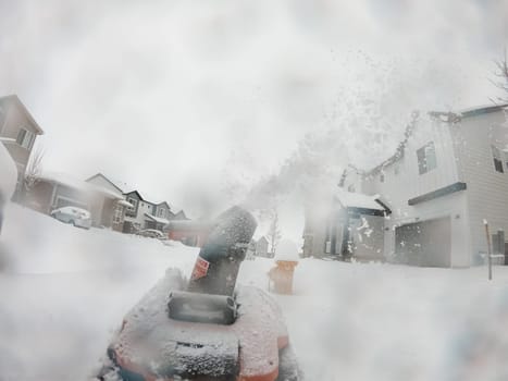 View from a snow-blanketed driveway captures a residential area mid-snowfall, with houses partially obscured by the cascading white flakes and a snow-covered car in the foreground.