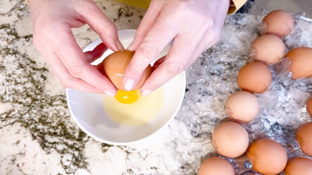 Hands are shown cracking a fresh egg open over a white plate, with a cluster of brown eggs waiting their turn on the cool, marbled surface of a well-used kitchen counter.