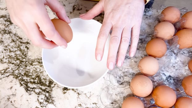 Hands are shown cracking a fresh egg open over a white plate, with a cluster of brown eggs waiting their turn on the cool, marbled surface of a well-used kitchen counter.
