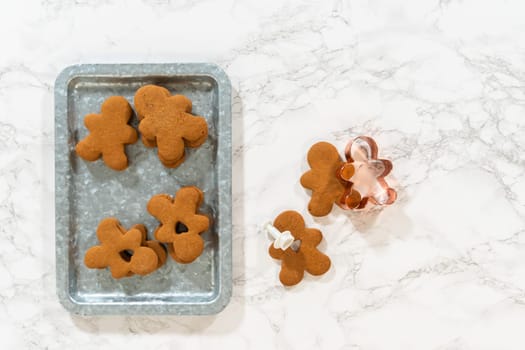 Flat lay. Gingerbread cookies, including a gingerbread man with a heart-shaped cutout, rest on a rustic metal tray against a marble countertop.