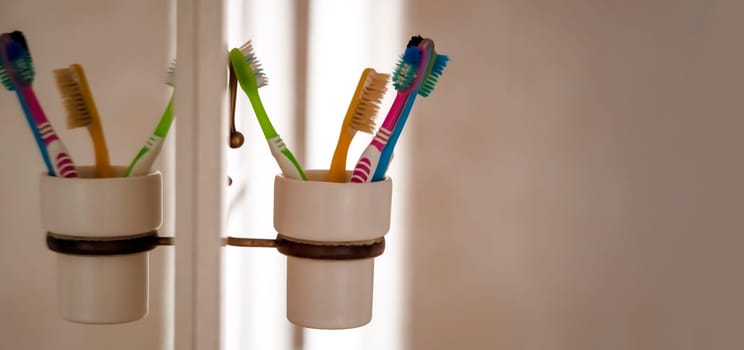 Toothbrushes in a glass attached to a mirror, personal hygiene products to take care of the cleanliness and health of the oral cavity, gums and teeth at home in the bathroom.