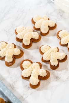 Gingerbread cookies await their second halves on a marble surface, each meticulously piped with buttercream to craft delightful sandwich treats. The precision of the piping adds a festive touch.