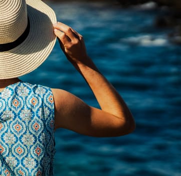 A young graceful girl in an elegant light blue dress with ornaments stands and looks at the endless ocean, a lady in a hat enjoys a vacation at sea, happily spends her leisure time during holidays.