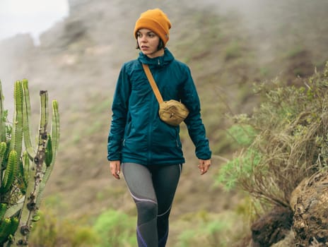 A young sporty girl in a windstopper jacket and thermal underwear walks along a path, hiking among the mountains in a national park with exotic vegetation, cacti and tropical plants.