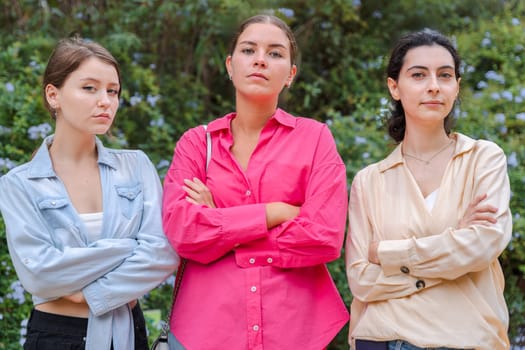 Three young Caucasian women stand outdoors with their arms crossed and a serious expression. They look confident and determined, posing together in front of the vegetation looking at camera.