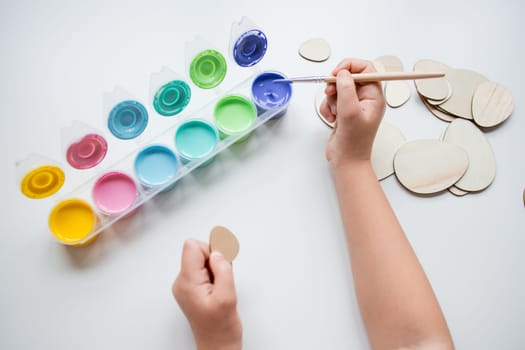 Child painting Easter wooden egg shapes with colorful paints on white table.