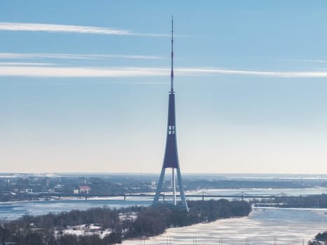The Riga TV Tower stands on an island in the Daugava River, surrounded by snow and frozen water, with Riga's cityscape and bridges in the background.