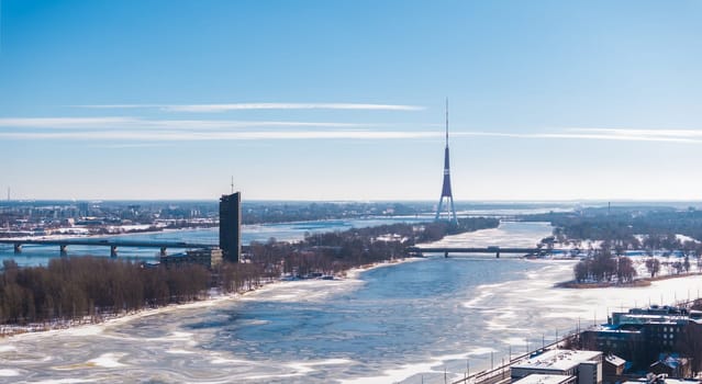Aerial view of Riga, Latvia, featuring the frozen Daugava River, Riga TV Tower, bridges, and the National Library amid a snow covered winter landscape.