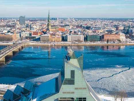 Aerial view of Riga, Latvia, featuring the National Library, icy Daugava River, St. Peter's Church spire, historic Old Town, and a snow covered landscape.