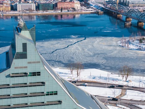 Aerial view of Riga, Latvia, featuring the National Library's angular design, the frozen Daugava River, a railway bridge, and snow covered old town buildings.
