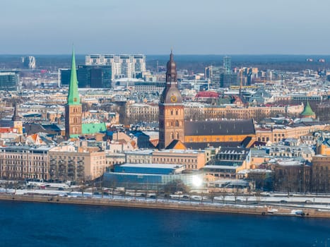 Aerial view of Riga in winter with the Daugava River, Riga Cathedral, St. Peter's Church, snow covered rooftops, and a mix of historic and modern buildings.