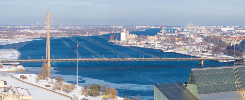 Aerial view of Riga, Latvia, in winter, showing the Vansu Bridge over the Daugava River, the National Library, a frozen river, and industrial structures.