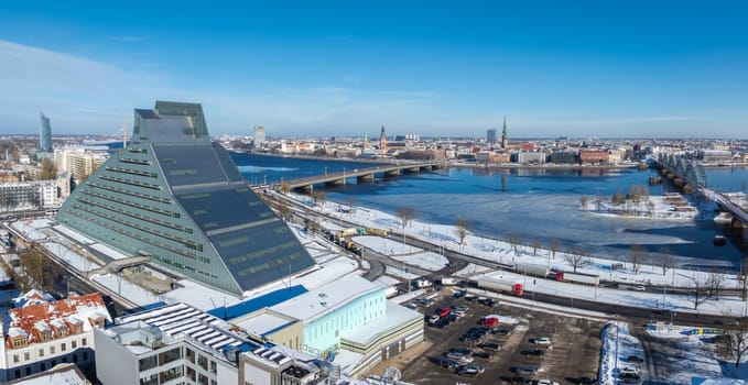 Aerial view of Riga, Latvia, showcasing the National Library's modern design, the Daugava River, snowy landscapes, and historic Old Town spires.