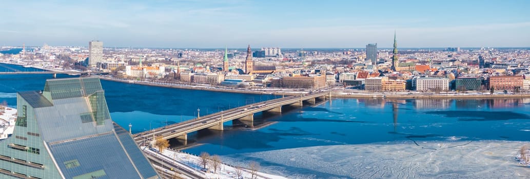 Aerial view of Riga, Latvia, in winter, featuring the Daugava River, National Library, Old Town landmarks, and a bridge under a clear blue sky.