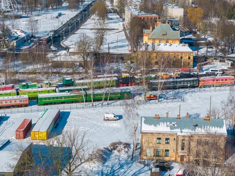 Snowy aerial view of Riga, Latvia, showcasing the Latvian Railway History Museum with colorful vintage train carriages, nearby buildings, and leafless trees.
