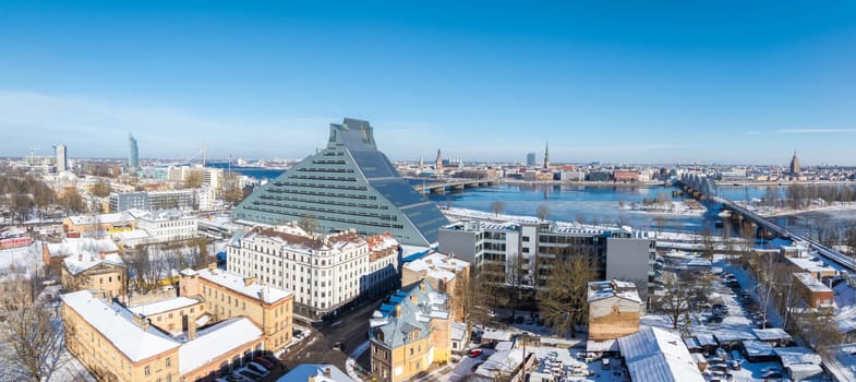 Aerial view of Riga, Latvia, featuring the National Library near the Daugava River, snow covered Old Town spires, TV tower, and railway bridge.