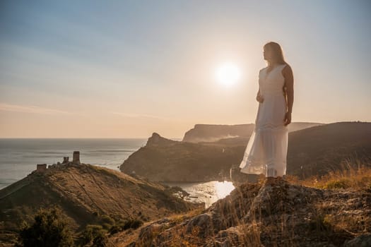 A woman stands on a rocky hill overlooking the ocean. She is wearing a white dress and she is enjoying the view. The scene is serene and peaceful, with the sun shining brightly in the background