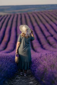 A woman wearing a straw hat stands in a field of purple flowers. She is holding a basket and she is enjoying the scenery