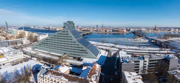 Aerial view of Riga, Latvia, in winter with the National Library, Daugava River, bridges, and historic Old Town spires under a clear blue sky.