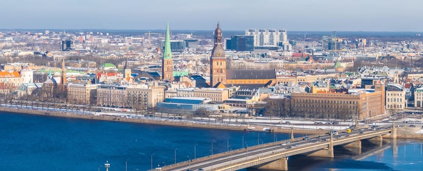 Aerial view of Riga, Latvia, in winter with the Daugava River, a bridge, Riga Cathedral, St. Peter's Church, historic old town, and modern buildings.