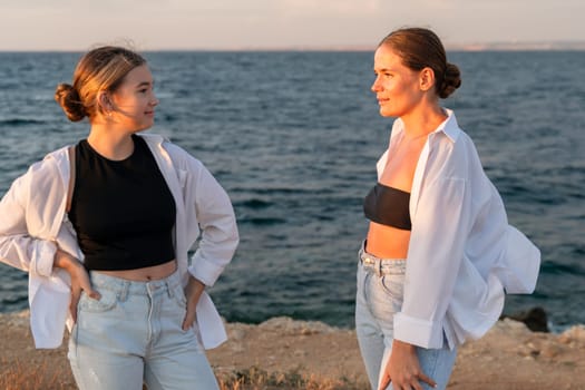 Two women, one in a white shirt and jeans, the other with a black top and white shirt tied around her waist, stand facing each other on a rocky shore against the backdrop of the ocean and a setting sun.