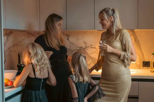 Two women and two girls gather in a modern kitchen. One woman holds a drink the girls are near a counter with fruit. They appear to be socializing.