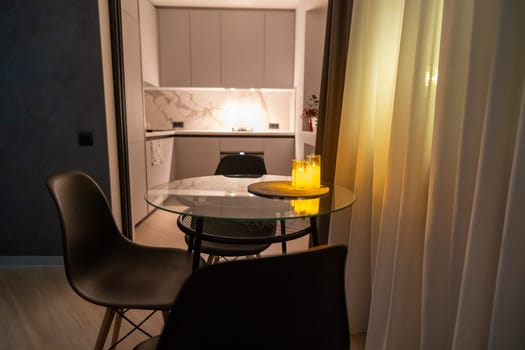 Modern apartment interior: glass-top table with two chairs, partially visible kitchen beyond, illuminated by warm lighting. Sheer curtains frame the scene.