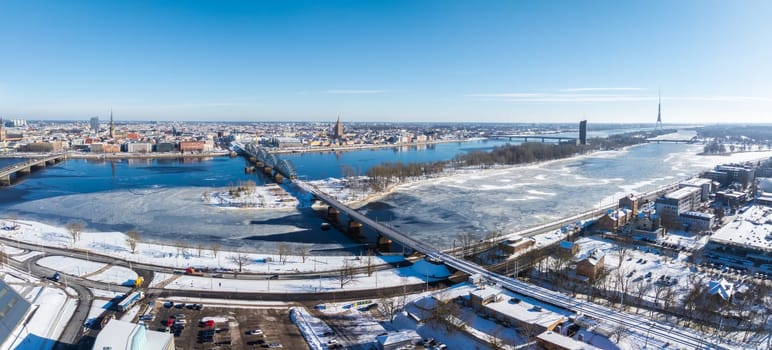 Aerial view of Riga, Latvia, in winter featuring the frozen Daugava River, National Library, Old Town spires, and Riga TV Tower under snow.