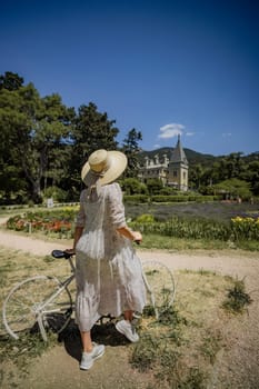 A woman in a white dress is standing next to a bicycle in a park. The scene is peaceful and serene, with a beautiful view of a house in the background. The woman is enjoying her time in the park