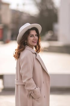 A woman wearing a white hat and a tan coat is standing in front of a building. She has a smile on her face and she is enjoying the moment