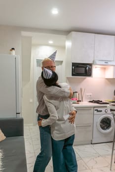 Here's a caption that describes the image accurately within the character limit: A man in a party hat hugs a woman in a kitchen. They embrace warmly. A washing machine and a sofa are visible in the background.