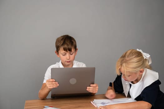 A boy and a girl, dressed in school uniforms, are working at a desk. The boy is looking at a laptop, while the girl is writing in a notebook.