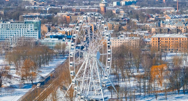 Aerial view of Riga, Latvia, in winter featuring a Ferris wheel, snow covered trees, a water tower, and a mix of historic and modern architecture.