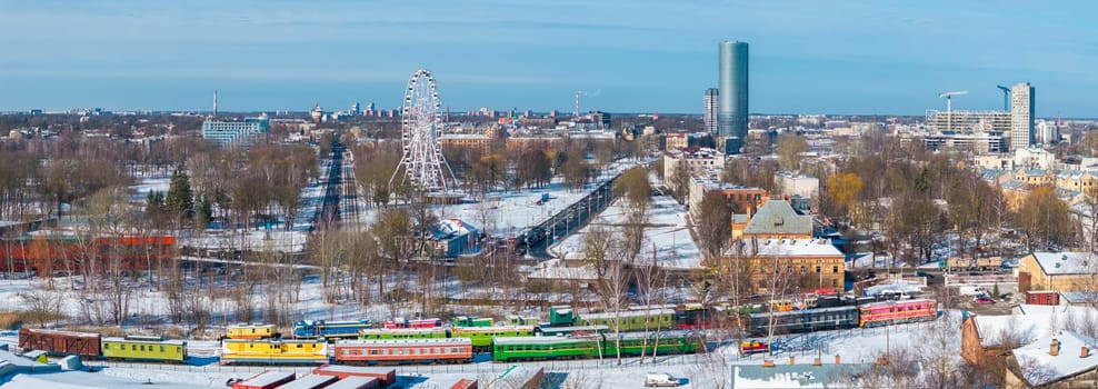 Snow covered Riga with colorful train carriages in the foreground, a Ferris wheel in the midground, and Z Towers among modern buildings in the background.