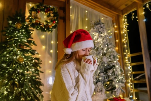 A woman wearing a Santa hat and white sweater holds a cup of coffee while standing in front of a decorated Christmas tree.