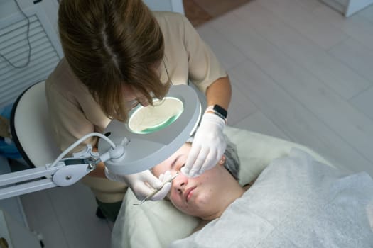 A cosmetologist uses a magnifying lamp to examine a client's face during a skin treatment. Close-up view shows hands gently working on the face.