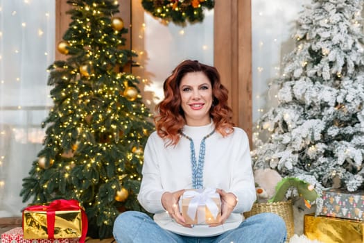 A woman with red hair sits in front of two decorated Christmas trees, holding a gift in her hands and smiling.