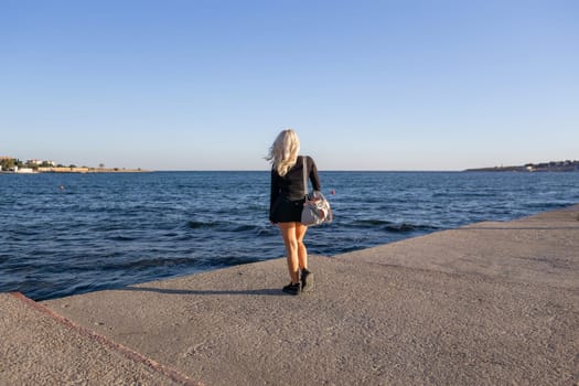 A woman with long blonde hair walks on a concrete pier overlooking a calm blue sea. She wears a black dress and carries a bag. The sky is clear and blue.
